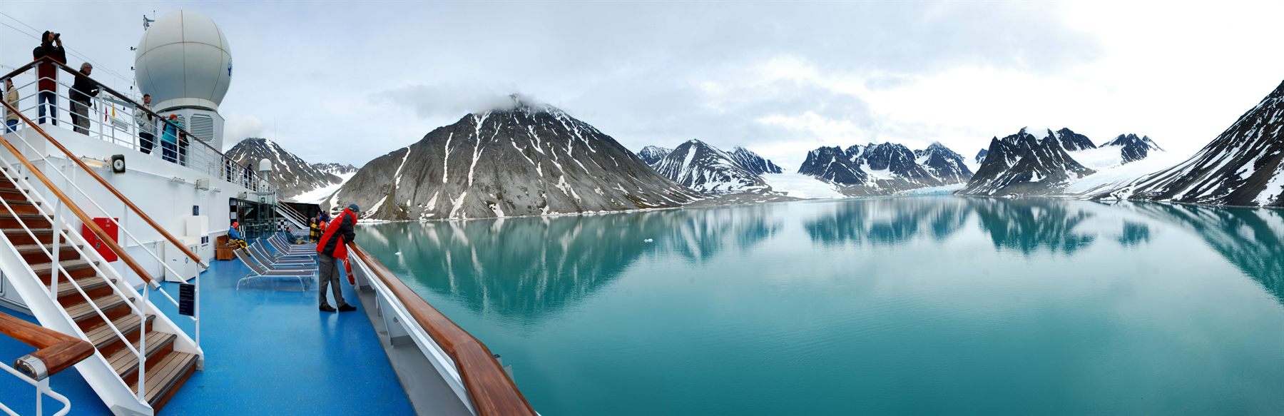 Passengers on the deck of an expedition ship in a calm fjord ringed by jagged snowy peaks